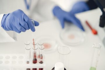 Blue-gloved hands using a pipette to transfer red liquid into glass test tubes on a lab bench