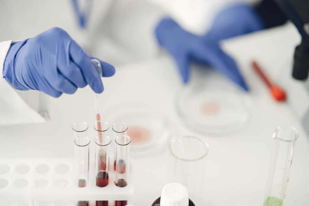 Blue-gloved hands using a pipette to transfer red liquid into glass test tubes on a lab bench
